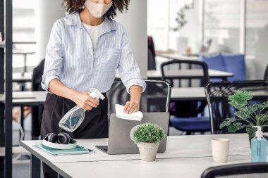 Koronavirüsü önleme. Koruyucu maskeli Afrikalı Amerikalı kız bilgisayarını sünger ve spreyle dezenfekte ediyor.