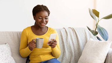 Yurtiçi rahatlama. Pozitif siyah kadın akıllı telefon ve kahveyle kanepede dinleniyor, internette geziniyor ve sıcak içeceklerin tadını çıkarıyor, Afrikalı kadın sosyal ağları kontrol ediyor veya arkadaşlarıyla mesajlaşıyor, boşluğu kopyalıyor.