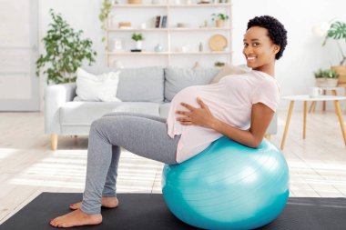 Hamilelik konseptinde egzersiz yapmak. Müstakbel pozitif anne evde fitness topu üzerinde çalışıyor. Bebek beklerken sağlıklı ve zinde kalan Afrikalı Amerikalı kadın bekleniyor.