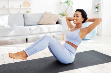 Ev ödevi konsepti. Spor kıyafetli güzel siyahi kadın içeride yoga minderiyle karın kası egzersizi yapıyor. Pozitif Afrikalı Amerikalı kadın karın kası egzersizi yapıyor, kaslarını güçlendiriyor.