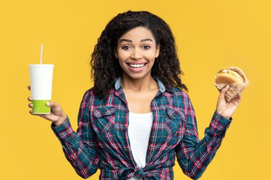 Fast Food Menüsü. Kıvırcık saçlı, gülümseyen Afro-Amerikan kadın portresi büyük lezzetli bir burger ve içecek tutuyor. Mutlu siyah kadın sandviçi yemeye hazır, turuncu arka planda izole edilmiş, afiş.