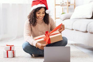Sanal Kutlama. Noel Baba şapkalı olumlu Afrikalı kadın kucağında Noel hediyesiyle oturuyor, oturma odasında hediye paketini açıyor, yerde oturuyor, dizüstü bilgisayarla video görüşmesi yapıyor.