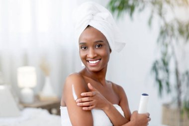 Güzellik bakımı, spa günü ve boş zaman. Havlu içinde gülümseyen Afrikalı Amerikalı genç bir kadına odaklan. Duş sonrası nemlendirici uygular. Modern yatak odasının iç kısmında bir şişe kozmetik tutar.