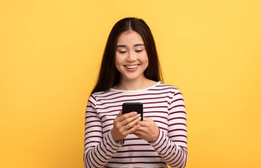 Akıllı telefonuyla etkileşime girerken çizgili uzun kollu üst üste sırıtan Asyalı genç bir kadın. Canlı sarı bir arkaplanın önünde duruyor, neşeli bir anı yakalıyor..