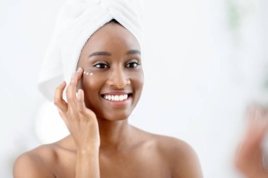 Neşeli genç bayan blogcu organik krem gösteriyor, sabahları duştan sonra yüzlerine uyguluyor. Kozmetik cilt bakımıyla tatmin olan Afrikalı Amerikalı kadın yatak odasındaki yumuşatıcı ürünü nemlendiriyor.