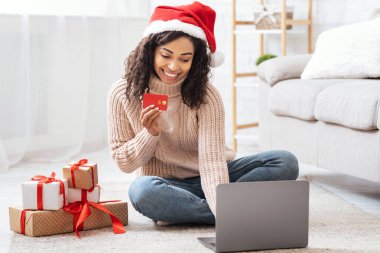 Kolay ödeme. Gülümseyen Afrikalı Amerikalı kadın Noel Baba şapkası takıyor, kırmızı kredi kartı gösteriyor, dizüstü bilgisayar kullanıyor hediye kutularının yanında oturuyor, yılbaşı ve yeni yıl hediyelerini online mağazada sipariş ediyor.