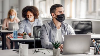Ofiste çalışmak ve yeni fikirler üretmek için yeni kurallar. Koruyucu maskeli Arap adam pencereden dışarı bakar ve iş yerindeki bilgisayarında antiseptik ve çiçekle çalışır.