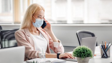 Yeni normallik ve karantinadan sonra işyerine dönmek. Koruyucu maskeli genç bir kadın akıllı telefonuyla konuşur ve laptopta çalışır, antiseptik ve bitkilerle aynı masada oturur.