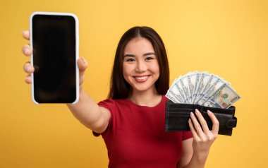 Neşeli genç bir kadın bir elinde akıllı bir telefon, diğer elinde nakit dolu bir cüzdan, parlak sarı bir arka plana gülümsüyor, finansal başarıyı ve modern teknolojiyi temsil ediyor..