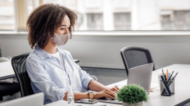 Yeni normal ve ofis işi koruyucu maske ve antiseptikle. Afrika kökenli Amerikalı bir kadın dizüstü bilgisayarında modern iç mekanlarda müşteri siparişleri yazmak için yazı yazıyor.