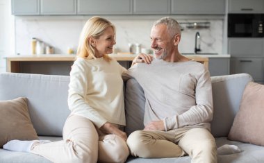 Neşeli bir çift, modern bir oturma odasındaki rahat bir kanepede rahatlar, açık renkli kıyafetler giyerken gülümsemelerini ve sevgilerini paylaşırlar. Sıcak atmosfer bağlantılarını güçlendiriyor..