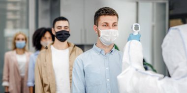 Virüs salgınına karşı önleyici önlemler. Koruyucu giysili adam, ofis içinde koruyucu maskeli işçilerin sıcaklığını ölçüyor, covid-19 'un yayılmasını engelliyor, serbest alan.