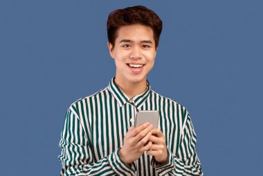 Harika bir uygulama. Akıllı telefon ayakta duran ve poz veren neşeli Asyalı genç adamın portresi. Mor stüdyo arka planında izole edilmiş, boş alan. Gülümseyen adam kameraya bakıyor, elinde aletle.