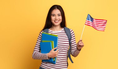 Neşeli bir öğrenci bir elinde Amerikan bayrağı, diğer elinde renkli ders kitaplarıyla, canlı sarı bir arkaplan önünde duruyor, okul ruhunu ve vatanseverliğini vurguluyor..