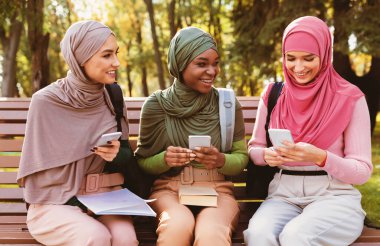 Parkta bankta oturan akıllı telefonlar kullanan üç modern Müslüman öğrenci. Sosyal medyada tesettürdeki Arap kadınlar telefonda mesajlaşıyor. Eğitim Uygulaması, Gadget Yaşam Biçimi