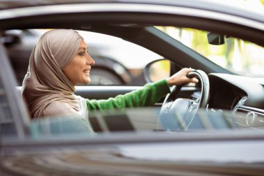 Kadın şöför. Yeni lüks arabasını kullanan başörtüsü takan İslamcı bir kadının yan görünüm portresi. tesettürlü gülümseyen müslüman kadın elinde direksiyon, dışarıdan bakıyor