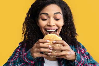 Abur cubur. Aç Afro-Amerikan Lezzetli Burger Yiyen Kadın portresi. Portakal Stüdyosu 'nun arka planında izole edilmiş, sağlıksız fast food' lara bakan komik bir kadın.
