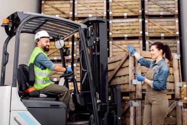 Elektrikli forkliftteki çalışan olgun elmalı konteynırı depolama kamerasına taşır, tabletli yönetici yönü gösterir. Üretim, ambalaj ürünleri depoda, modern aletler hasat için.
