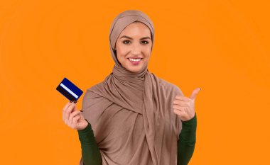 Sarı Stüdyo Arkaplanının Üzerinde Duran Kameraya Gülümseyen ve Bankacılık Hizmetlerini Onaylayan Başparmağıyla Kredi Kartı Tutan Gülümseyen Arap Kadın. Banka Konseptimi Seviyorum