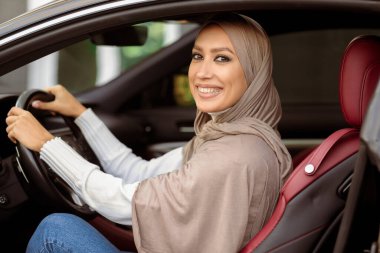 Şehir turu. Türbanlı Mutlu Müslüman Kadın Modern Araba Kullanıyor, Direksiyon Tutuyor 'un yan görünümü. Arap kadın yeni otomobil için test sürüşü yapıyor, kameraya bakıyor ve poz veriyor