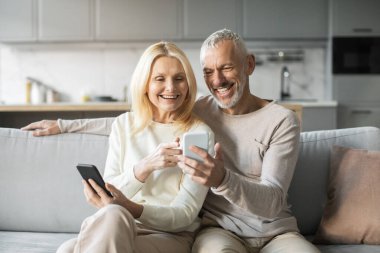 Yaşlı bir çift rahat bir koltukta oturur, gülümser ve akıllı telefonlarıyla meşgul oldukları bir anı paylaşırlar. Sıcak atmosfer onların neşeli etkileşimlerini vurgular..