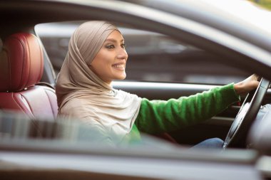 Kendine güvenen sürücü. tesettürlü gülümseyen müslüman kadın modern araba kullanıyor. Türbanlı genç bir kadın yeni bir araca biniyor, direksiyon tutuyor ve yola bakıyor.