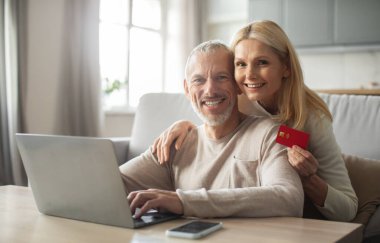 Neşeli bir son sınıf çifti bir masada birlikte oturuyor, bir eşi dizüstü bilgisayar kullanırken diğeri de kredi kartı tutuyor, hem gülümsüyor hem de rahat yaşam alanlarında keyifli bir anı paylaşıyor..