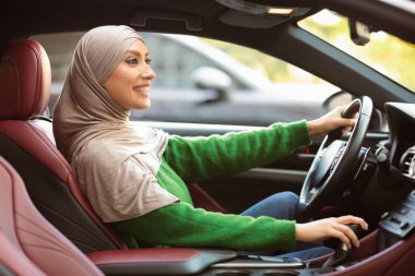 Test sürüşü konsepti. Kentte mutlu Müslüman bir kadının araba sürüşünün, yeni otomobilini kontrol edişinin, araç alışının, sürücü koltuğunda oturuşunun, direksiyon tutuşunun ve vites kolunun yan görüntüsü.