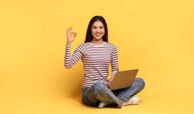 Gülümseyen genç bir kadın dizüstü bilgisayarını tutarken sarı bir zeminde bacak bacak üstüne atmış oturuyor. Her şeyin yolunda olduğunu göstermek için eliyle el hareketi yapıyor. Kendine güven ve pozitiflik yayıyor..