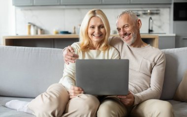 Üst düzey bir çift rahat bir kanepede rahatlar, güzel tasarlanmış oturma odalarında dizüstü bilgisayara bakarken kahkahalarını paylaşırlar. Sıcak atmosfer, mutluluklarını ve bağlantılarını vurgular..
