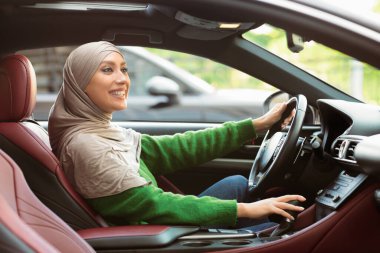 Şehirde at sürmek. Modern araçta oturan mutlu Müslüman kadın, yeni arabasını kullanıyor, direksiyon ve vites kollarında. Deneyimli kadın sürücü, yolculuğunun ve yolculuğunun keyfini çıkarıyor.