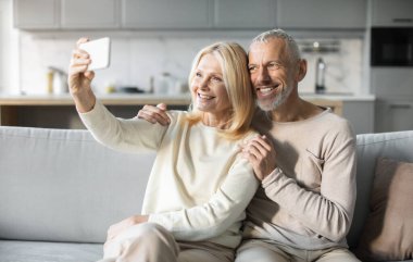 Neşeli bir son sınıf çifti rahat bir kanepede oturup selfie çekerken gülümserler. Güneş ışığıyla dolu, parlak, hoş geldin oturma odalarında sıcak bir anı paylaşıyorlar..