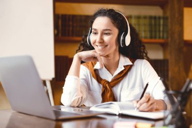 Ödev yapıyorum. Masada oturan gülümseyen genç bir kadının portresi kalemle, yazarak ve not alarak, dizüstü bilgisayarını kullanarak ve bakarak. Neşeli kıvırcık bayan çevrimiçi ders izliyor.