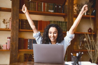 Evet, ben bir kazananım. Heyecanlı duygusal mest olmuş bir bayan, başarıyı, zaferi ya da harika haberleri kutluyor, masada oturuyor, kişisel bilgisayarını kullanıyor, evet diye bağırıyor ve ellerini kaldırıyor. Gülümseyen mutlu kadın neşe içinde