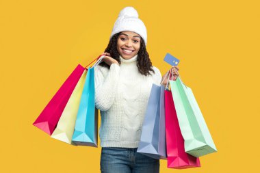 Kolay alışveriş konsepti. Kış şapkalı, kazaklı, kredi kartlı, alışveriş merkezinden gelen parlak renkli çantalı, turuncu arka planda kapalı duran mutlu siyah kadın. Perakende ve Satın Al