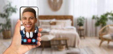 Bir kişi video görüşmesi sırasında ekranda gülümseyen bir yüz gösteren bir akıllı telefon tutuyor. Bu sahne, arka planda bitkilerin ve rahat mobilyaların bulunduğu şık bir evde geçiyor..