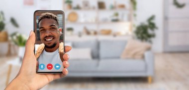 Bir insan rahat bir oturma odasında otururken akıllı bir telefonla neşeli bir görüntülü konuşma yapıyor. Arka planda rahat bir kanepe ve sıcak bir atmosfer yaratan bitkiler var..