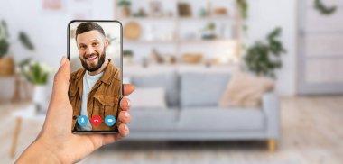 Video görüşmesi sırasında bir kişi akıllı telefon tutuyor. Arka planda kanepesi, bitkileri ve dekoratif rafları olan rahat bir oturma odası var. Rahat bir atmosfer yaratıyor..