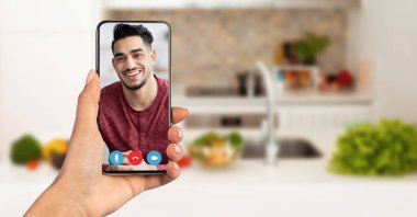 Bir kişi video görüşmesinde bir arkadaşıyla bağlantı kurmak için akıllı telefon kullanıyor. Arka planda taze sebzelerle dolu, sıcak bir ev atmosferi yaratan modern bir mutfak yer alıyor..