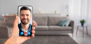 Bir kişi video görüşmesi sırasında gülümseyen bir kişiyi gösteren bir akıllı telefon tutuyor. Dekorasyon, modern telekomünikasyonun göstergesi olarak kanepesi olan rahat bir oturma odası..