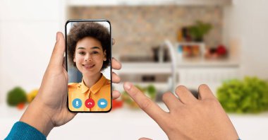 Görüntülü konuşma sırasında ekranda gülümseyen birini gösteren bir akıllı telefonu tutan bir el. Arka planda taze sebzelerle dolu parlak bir mutfak görünüyor, canlı bir atmosfer yaratıyor..