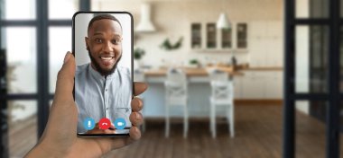 Bir insan mutlu bir şekilde akıllı telefonuyla çevrimiçi bir iletişim kuruyor ve dost canlısı bir atmosfer sergiliyor. Arka planda şık dekorları ve mobilyaları olan rahat bir mutfak yer alıyor..