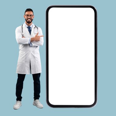 Gözlüklü Orta Doğulu erkek doktor ve mavi stüdyonun arka planındaki büyük telefon ekranını gösteren beyaz üniformalı doktor. Genç Doktor cep telefonunun yanında duruyor. Dikey Mockup Banner. Teletıp