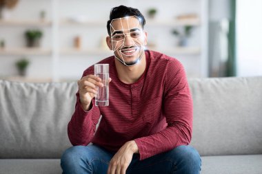 Bir adam bir bardak su tutarken gülümseyerek koltuğa rahatça oturur. Yüz hatları tanımlama teknolojisiyle vurgulanır, modern gelişmeleri tanımlamada sergiler..