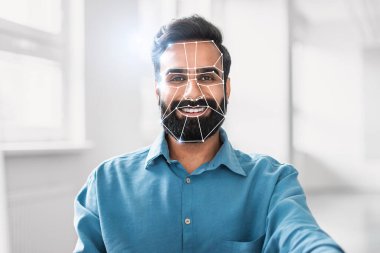 Hintli adam, yüzü, parlak ve modern bir ofiste yüz tanıma teknolojisi tarafından taranırken gülümsüyor. Bu, günlük faaliyetlerde dijital kimliğin giderek artan kullanımını temsil etmektedir..