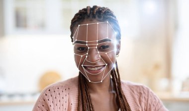 Afrika kökenli Amerikalı genç bir kadın, yüz tanıma teknolojisi onu kapalı bir ortamda tanımlarken gülümsüyor. Bu sahne, modern tekniklerin kimlik tespiti için artan kullanımını vurgular.