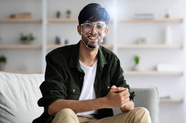 Genç bir Hintli adam koltukta rahatça oturuyor, yüz tanıma teknolojisi onun özelliklerini incelerken gülümsüyor. Bu, modern teknolojinin günlük tanımlama süreçlerindeki rolünü vurgular.