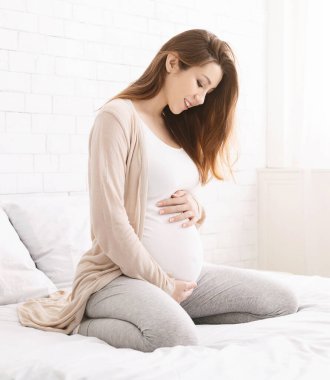 Gebelik, dinlenme, insanlar ve beklenti. Yatağın üstüne oturan ve karnı yavaşça evde dokunmadan mutlu hamile kadın