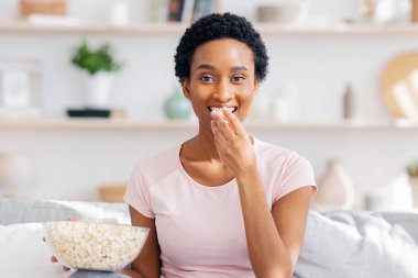 Akılsız yeme konsepti. Genç siyahi kadın evdeki rahat kanepede televizyon karşısında patlamış mısır yiyor. Afrikalı Amerikalı kadın film izlerken ağzını abur cuburla dolduruyor.