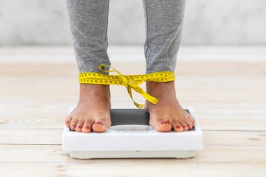 Tanınmayan siyah kadın terazinin üzerinde duruyor, ağırlığını bacakları şeritle bağlanmış şekilde kontrol ediyor. Yeme bozukluğundan muzdarip genç bir bayanın zayıf düşürme fikrine saplantılı görüntüsü.